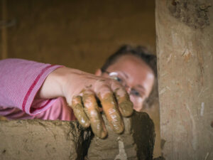 Eine Person mauert mit Lehmsteinen und hat sehr matschige Hände. Am Bildrand ist ein alter Holzbalken zu sehen.